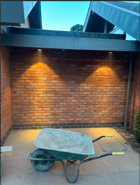 Freshly laid red brick feature wall lit by warm downlights in a private courtyard, with a builder's wheelbarrow in the foreground